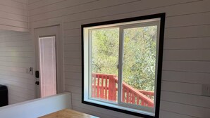 A room with a wooden bench and a table, a window with a view of a red deck and greenery.