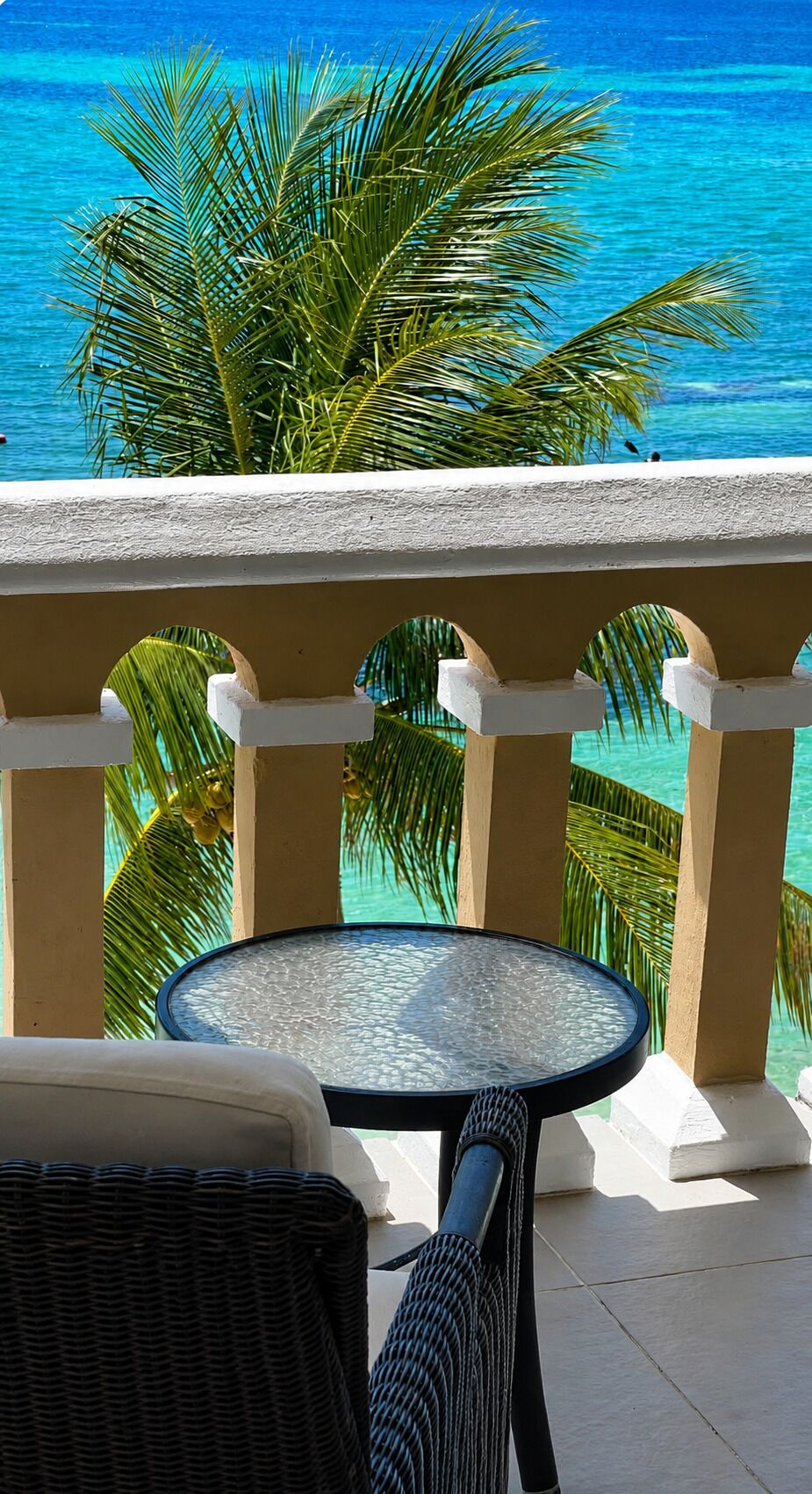 A balcony with a round table and chairs, overlooking a turquoise ocean with palm trees.