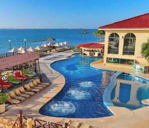 A swimming pool with a curved design, lounge chairs, and umbrellas, with a view of the sea and a building with a red roof. - caribbean apartments (Cancún)