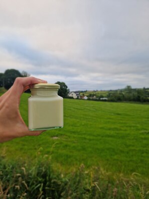 A hand holding a small glass jar with a white substance, set against a backdrop of a green field and a cloudy sky.