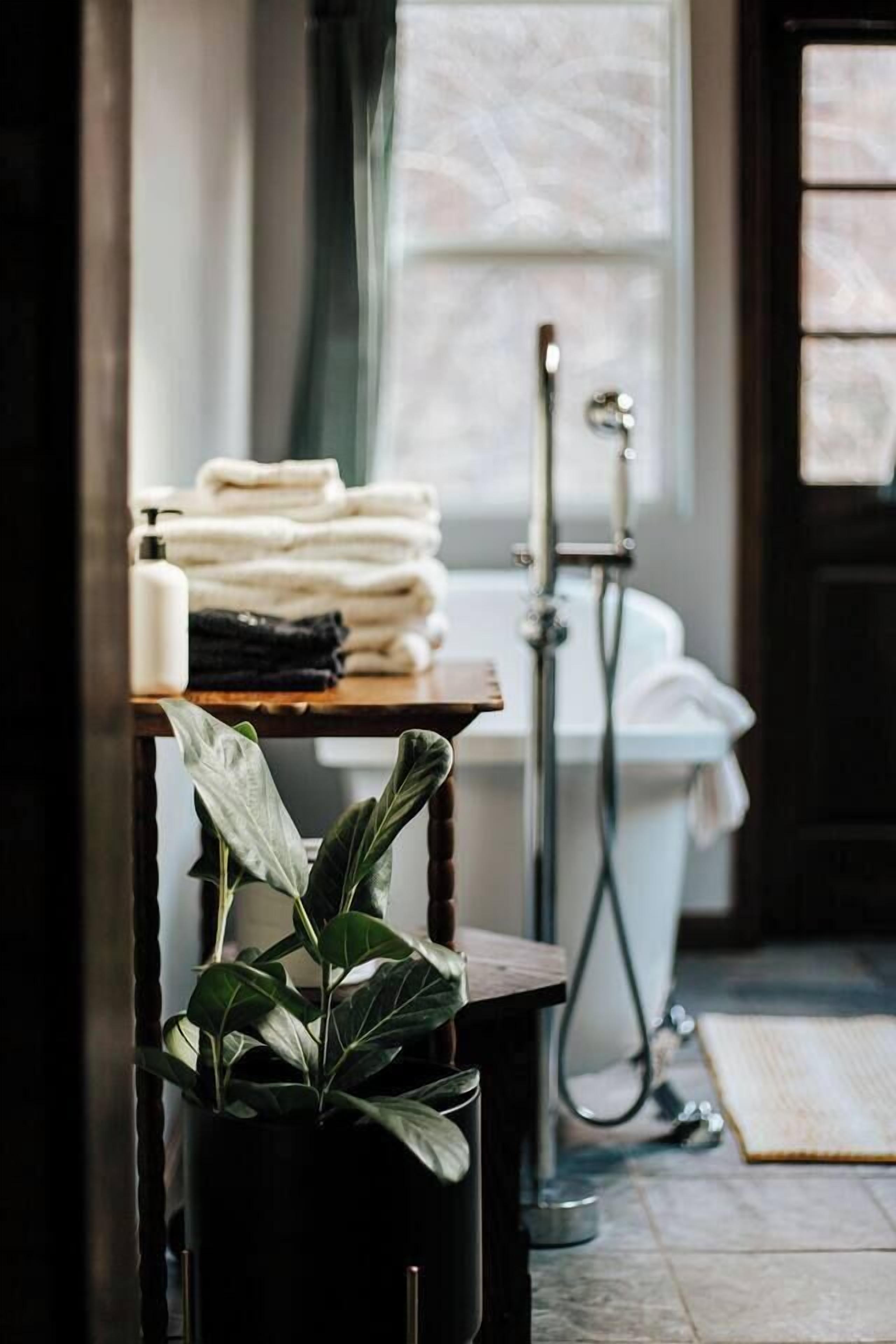 A bathroom with a bathtub, a towel rack with folded towels, a potted plant, and a window with curtains.