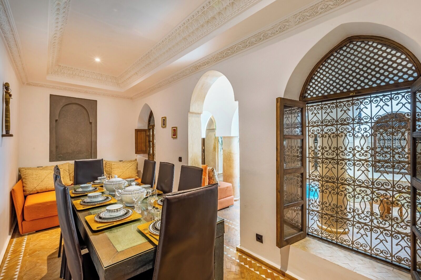Un coin repas avec une table dressée pour quatre personnes, entourée de chaises, et une vue sur une cour intérieure, accessible par une porte ouverte ornée de motifs en métal.