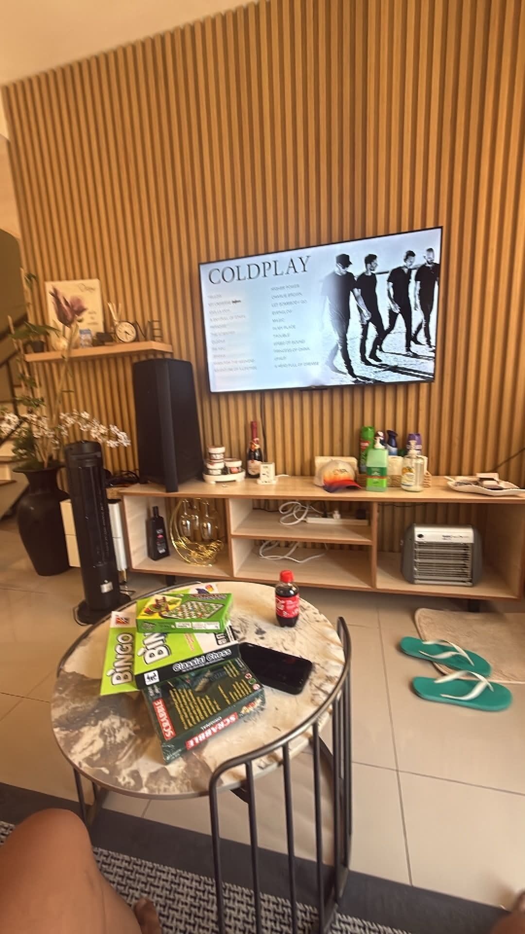 A living room with a television displaying a music album cover, a small round table with board games, a pair of flip-flops, and a speaker on a wooden shelf.