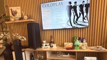 A living room with a television displaying a music album cover, a small round table with board games, a pair of flip-flops, and a speaker on a wooden shelf.