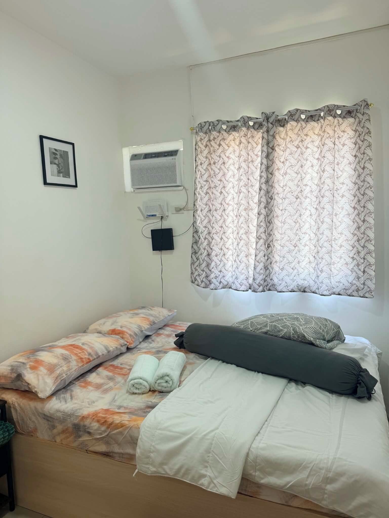 A single bed with a patterned bedspread, a rolled towel, and a pillow. An air conditioning unit is mounted on the wall, and a curtain is partially drawn.