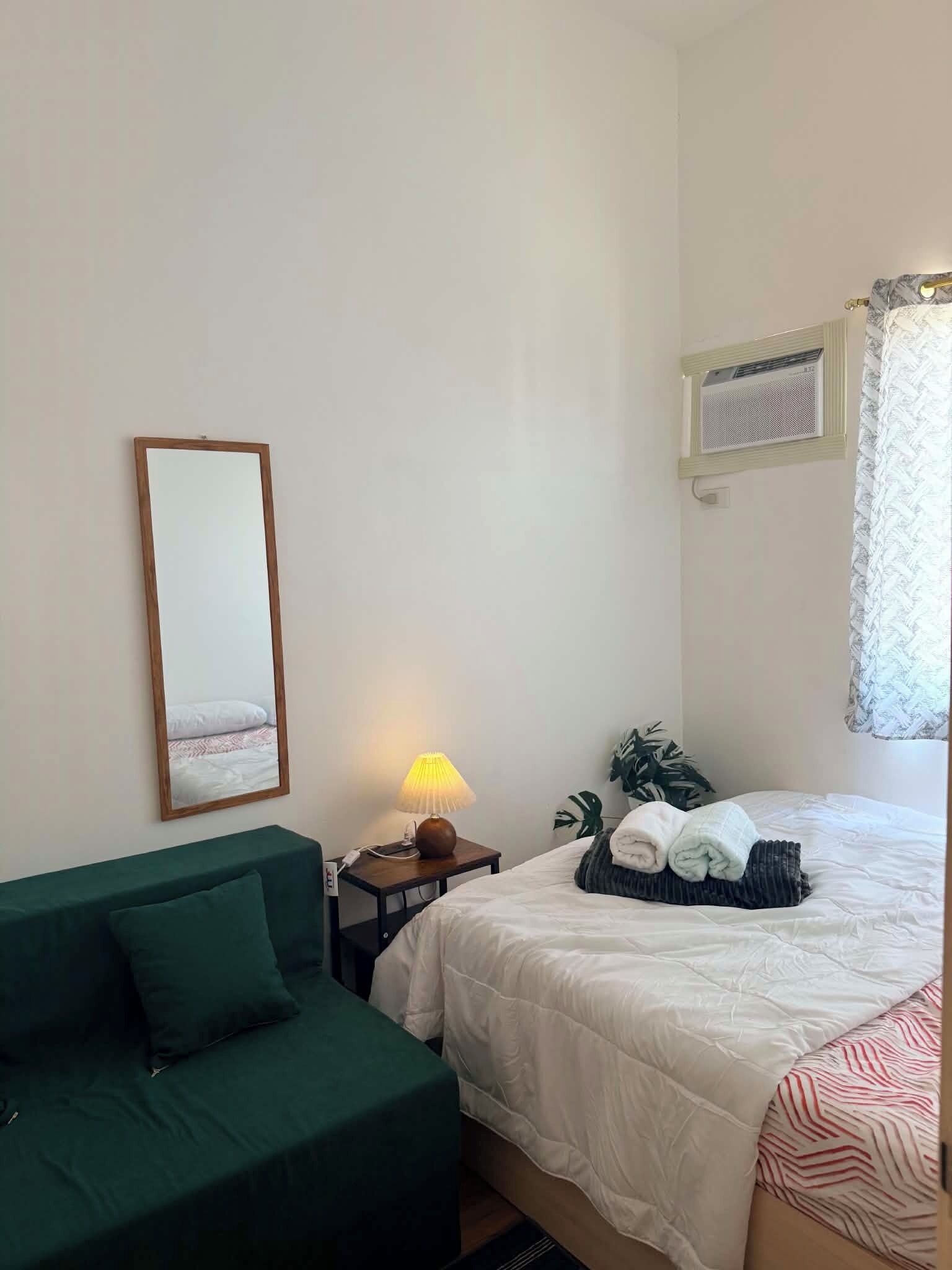 A hotel room with a bed, a green sofa, a bedside table with a lamp, a mirror, and a window with a curtain.