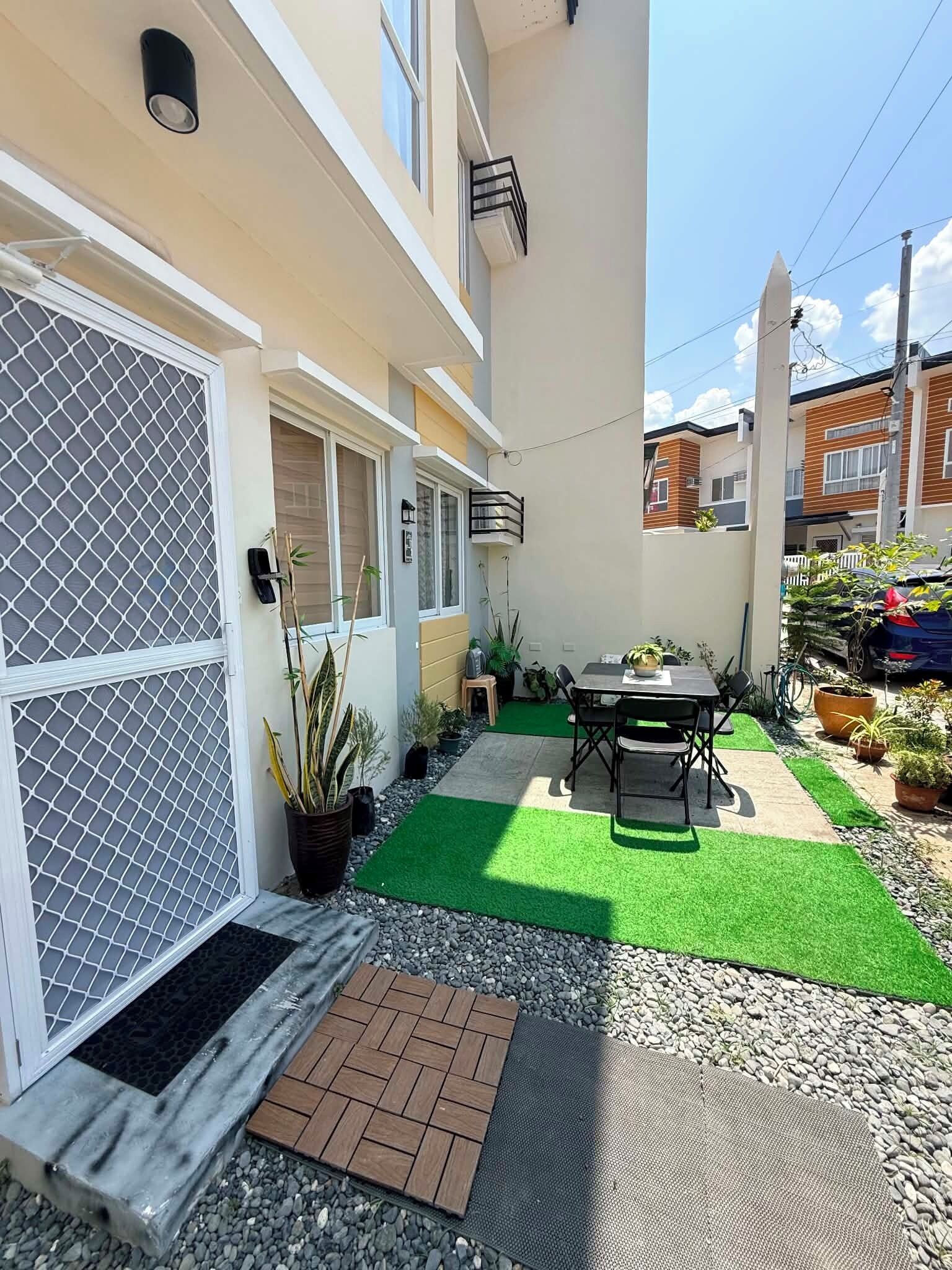 A residential outdoor space with a patio, a table with chairs, potted plants, and a green artificial turf area.