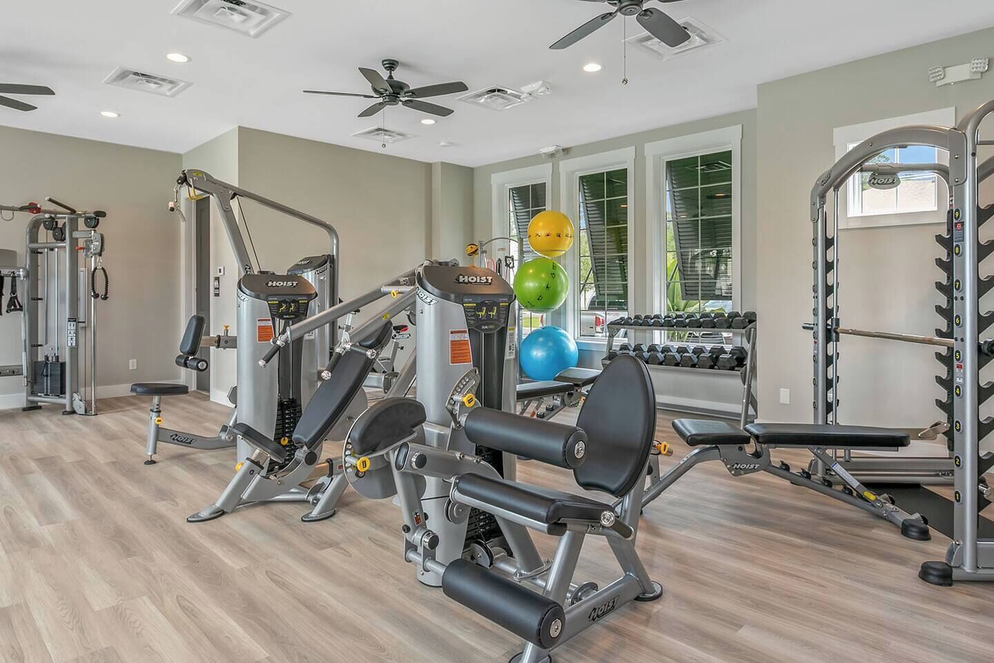 Ein Fitnessraum mit unterschiedlichen Trainingsgeräten, einem Deckenventilator und großen Fenstern.