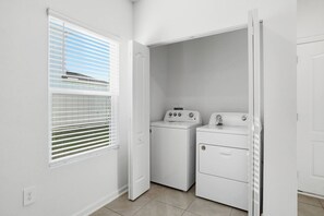 A laundry room with a washing machine and dryer, a window with blinds, and a door leading to another room.