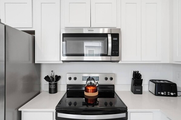 A kitchen with a stainless steel microwave, a gas stove with a kettle, a toaster, and a utensil holder.