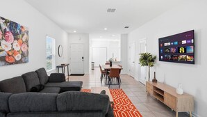 A modern living room with a sofa, a dining area with chairs and a table, and a TV mounted on the wall displaying various channels. - Posh 3BR Townhome| Pool | Gym | Close to Beach (St. Augustine)