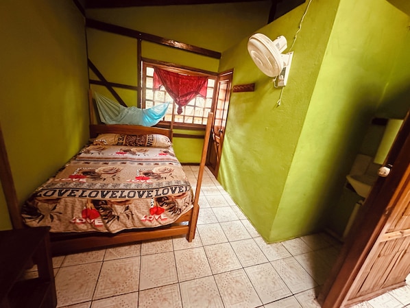 A small, single-bed room with a wooden frame, a patterned bedspread, a window with a red curtain, a wall-mounted fan, and a green wall.