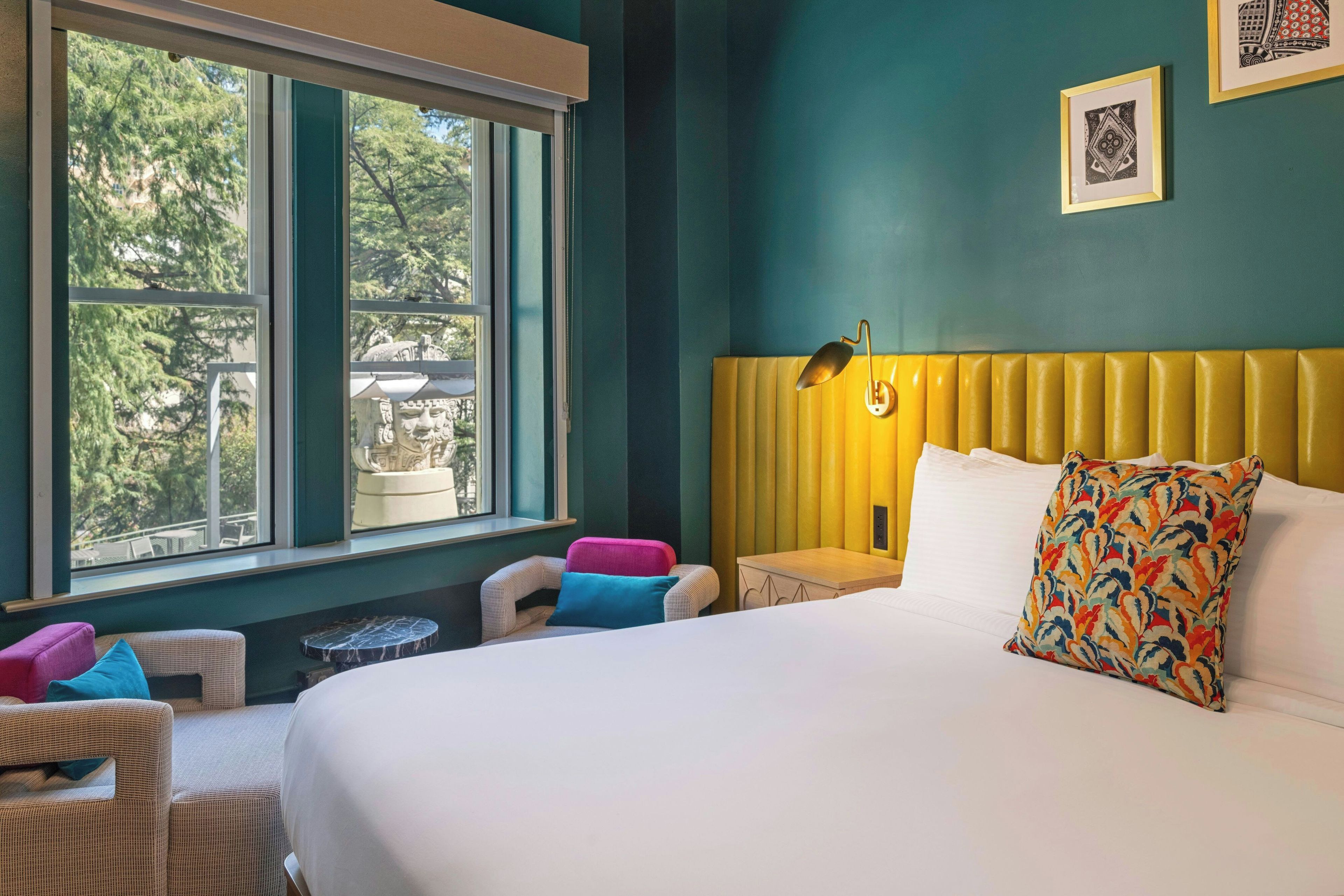 Habitación de hotel con una cama grande, una ventana con vistas a árboles, una mesita pequeña y una silla.