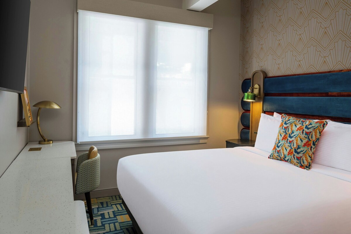 A hotel room with a large bed, a desk with a lamp, a chair, and a window with blinds.