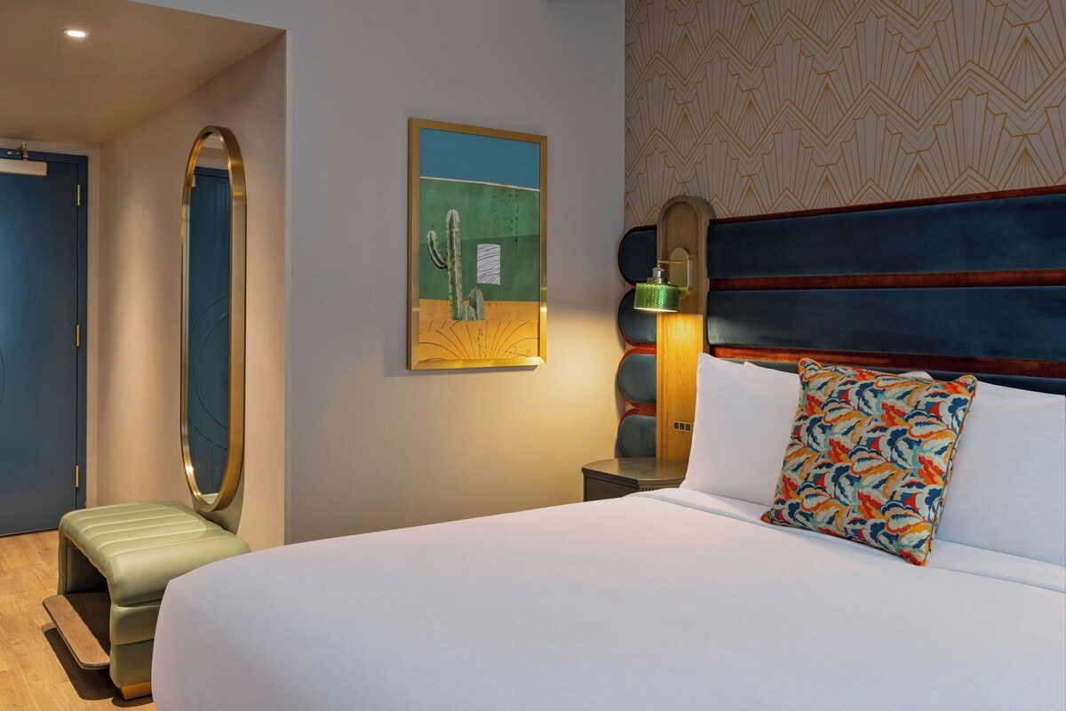 A hotel room with a bed, a nightstand, a lamp, a mirror, and a painting on the wall.