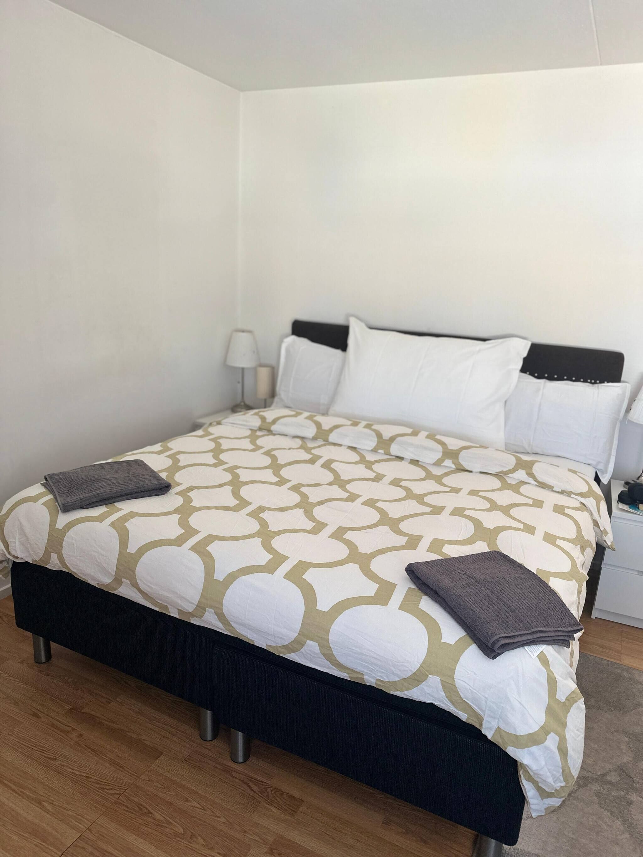 Ein ordentlich gemachtes Bett mit einem gemusterten Überwurf, ein Nachttisch mit Lampe sowie ein weiterer Nachttisch ebenfalls mit Lampe.