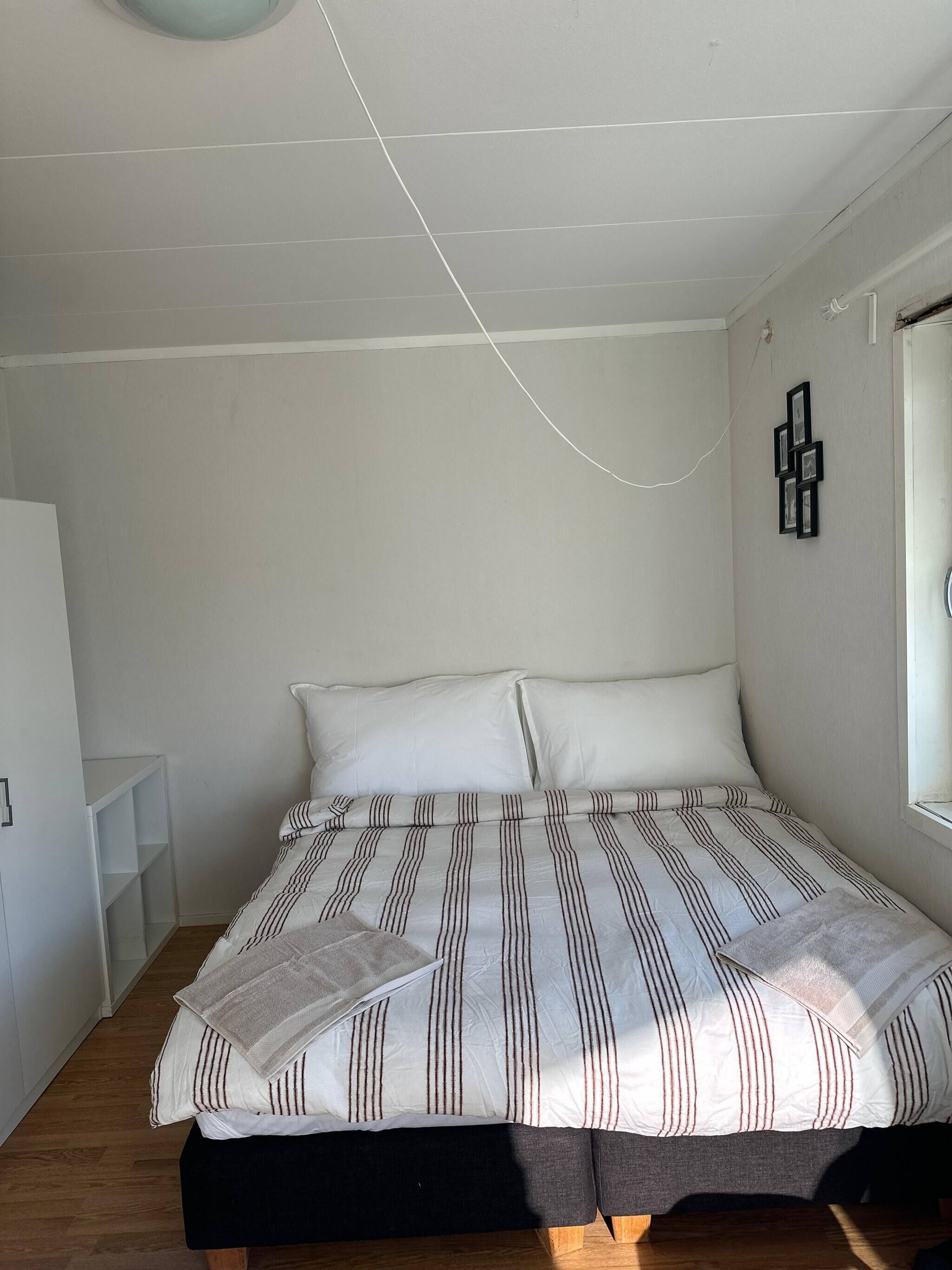 A bedroom with a bed, a white wall, a window, and a small white shelf.
