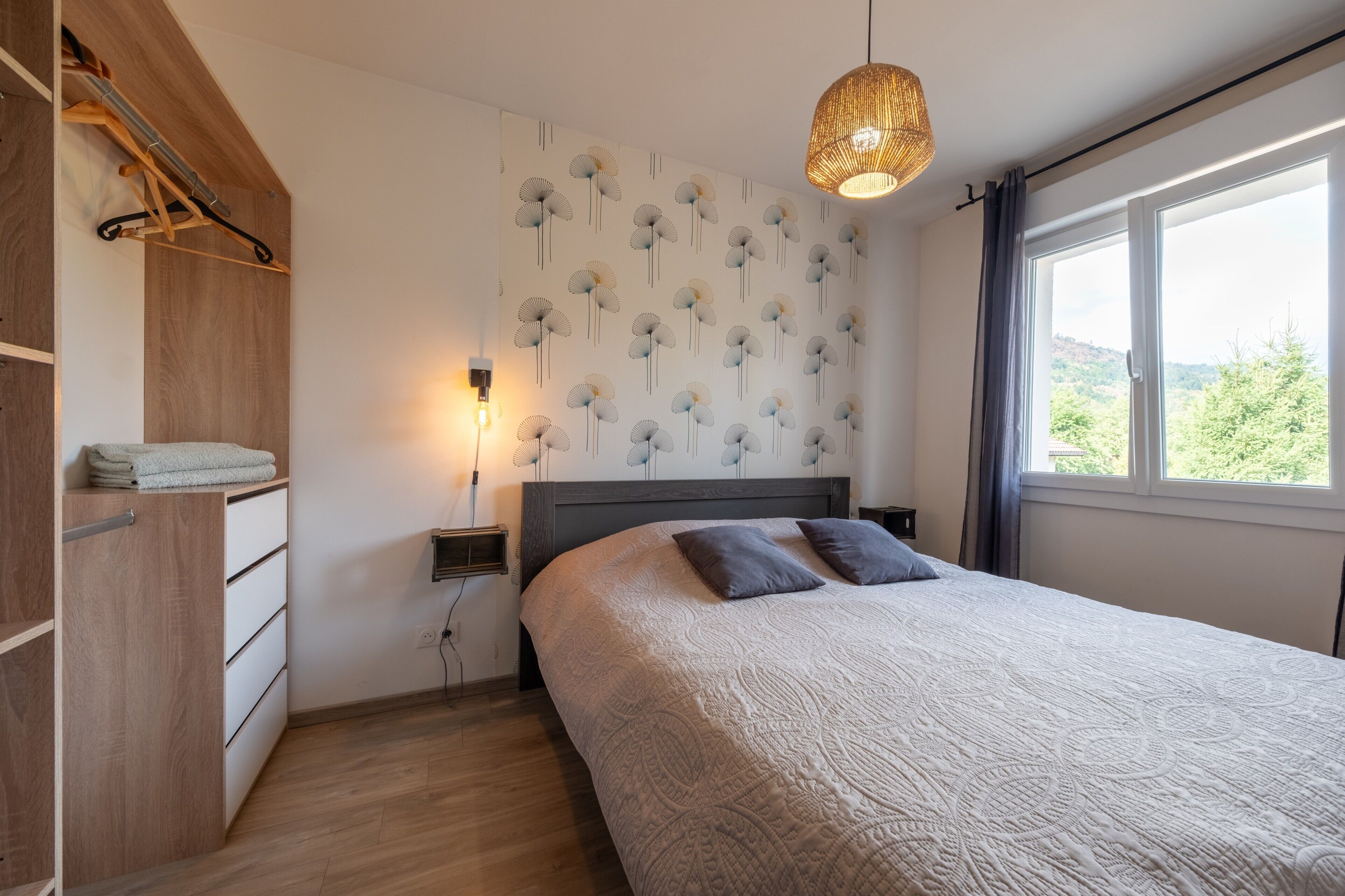 Une chambre à coucher comprenant un lit, une table de chevet, une lampe fixée au mur et une fenêtre donnant sur un paysage verdoyant.