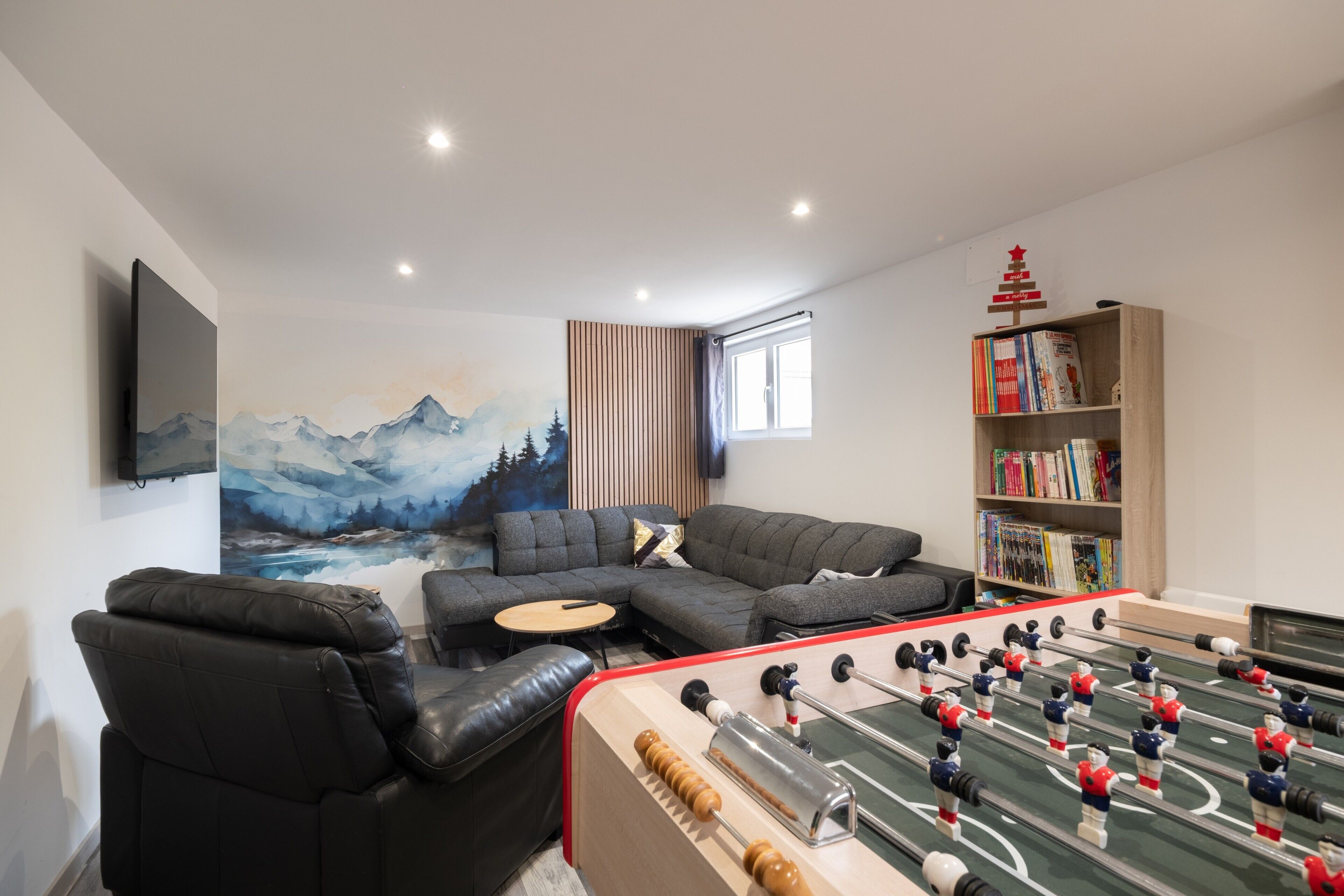 A modern living room with a foosball table, a sofa, a bookshelf, and a painting of mountains.
