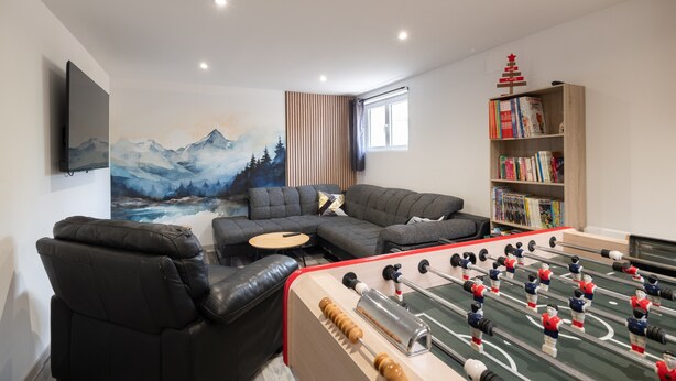 A modern living room with a foosball table, a sofa, a bookshelf, and a painting of mountains.