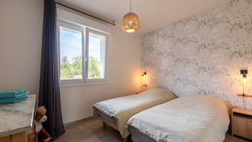 Ein Schlafzimmer mit zwei Betten, einem fensterbelaubten Fenster, einer Wandleuchte und einem Nachttisch aus Holz.