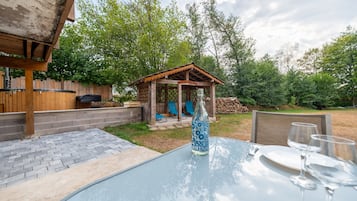 A backyard with a glass table, chairs, a pergola, and a wooden gazebo.