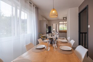 Un espace repas avec une longue table en bois dressée pour un repas, entourée de chaises, et un luminaire suspendu au-dessus.