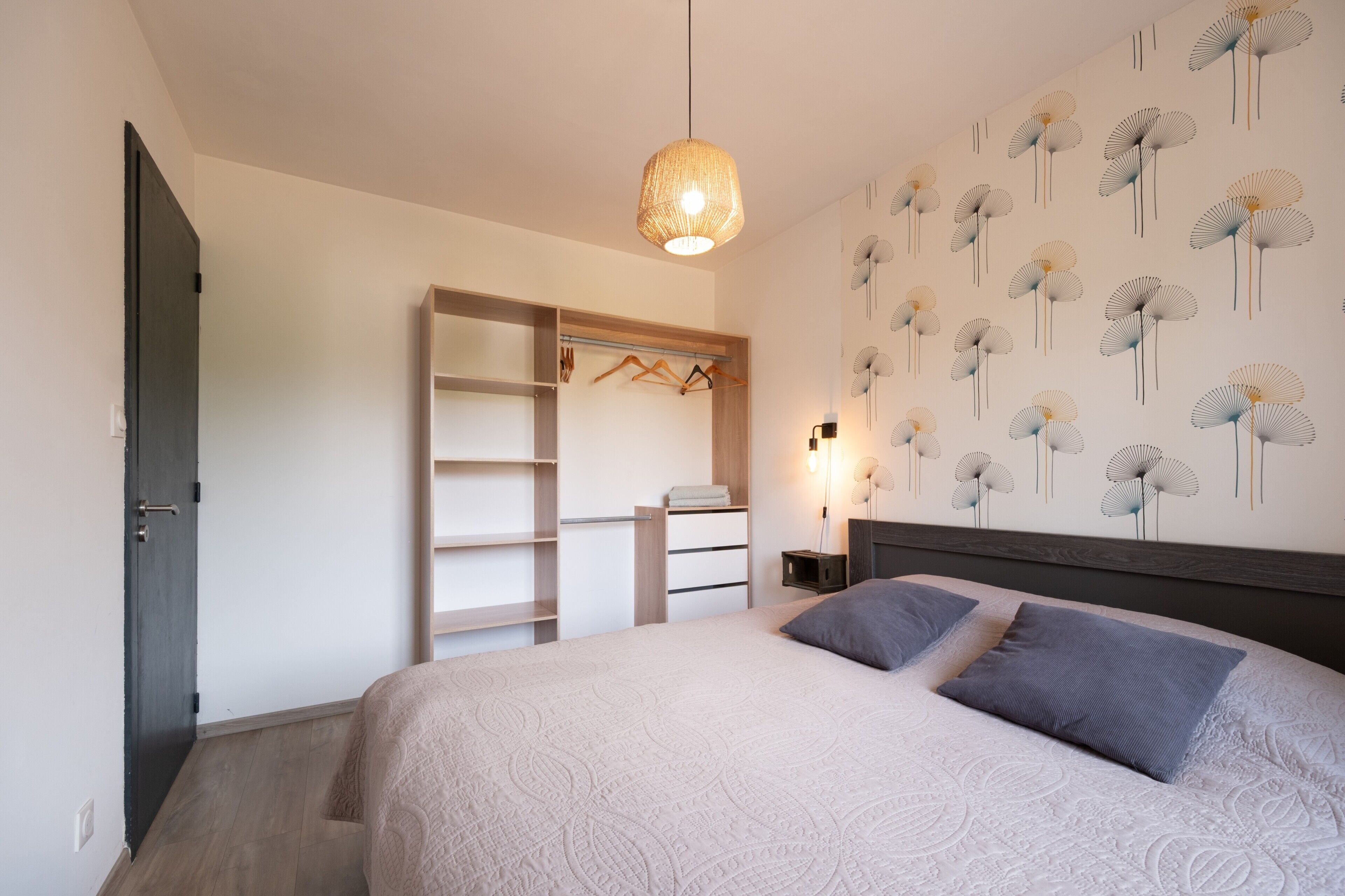 A bedroom with a bed, a hanging light fixture, a wardrobe, and a wall with a floral pattern.