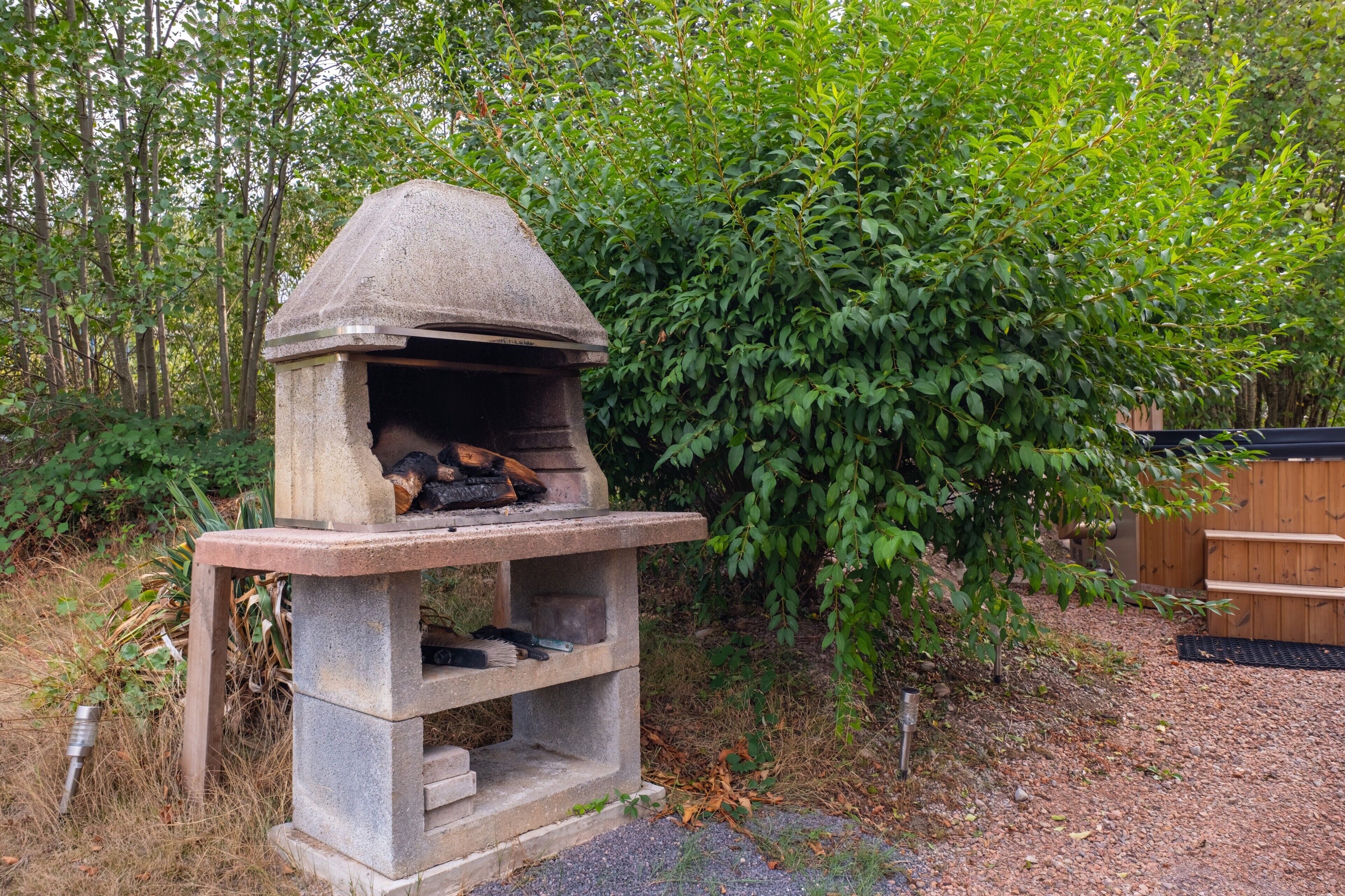 Un horno de piedra al aire libre con fuego encendido, rodeado de vegetación y una valla de madera.