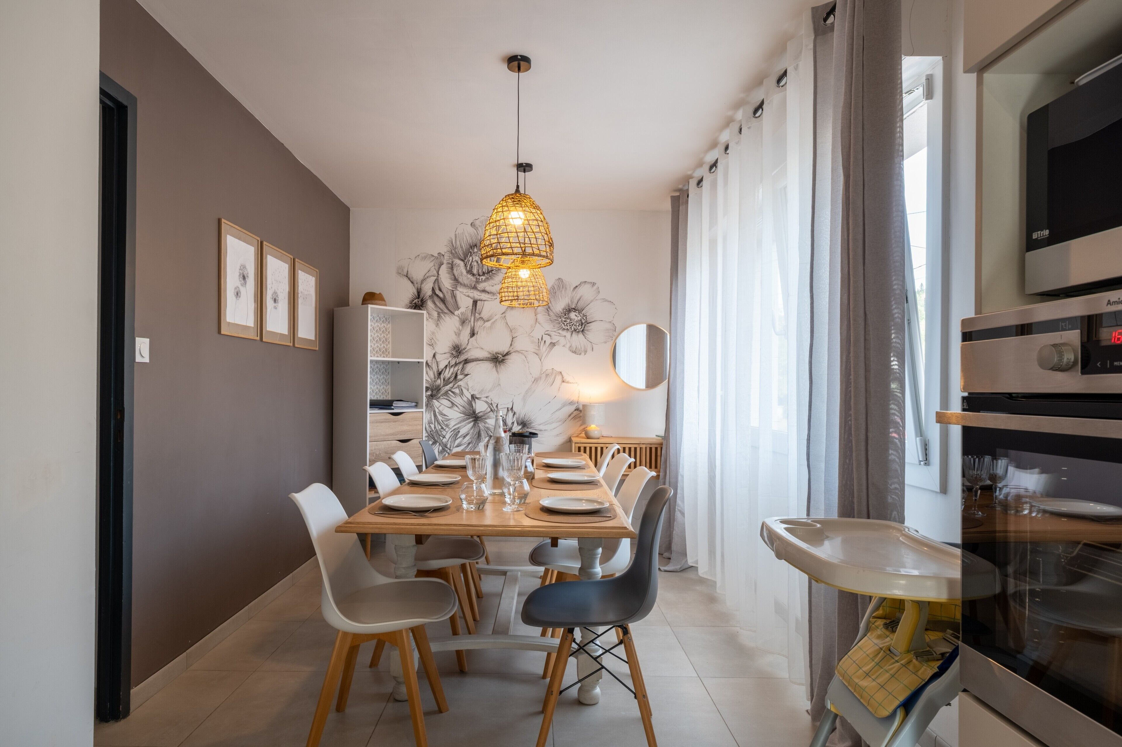 Un espace repas moderne avec une table en bois dressée pour quatre personnes, entourée de chaises, d’un luminaire suspendu et d’un mur orné d’une fresque florale.