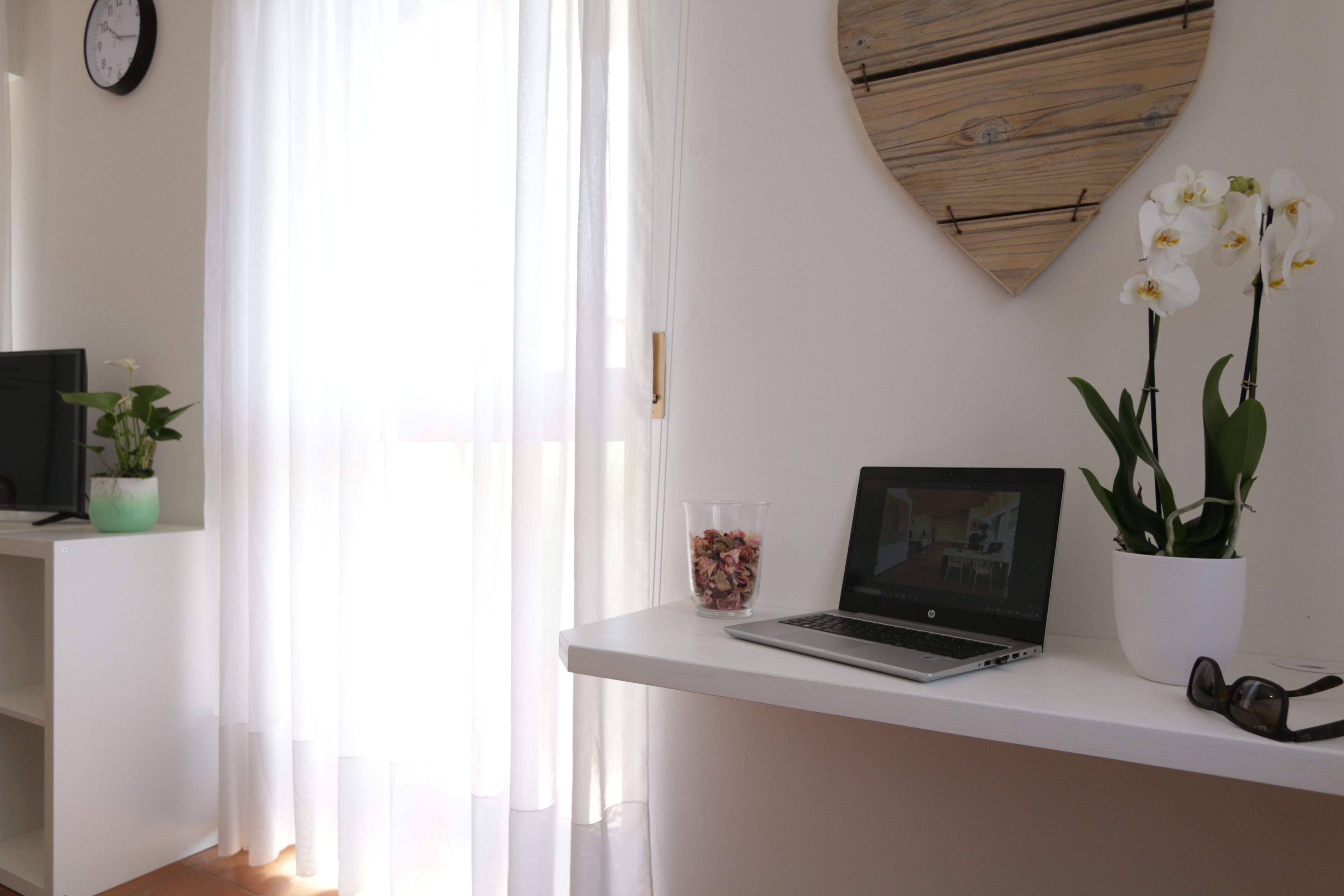 Ein Zimmer mit einem weißen Schreibtisch, einem Laptop, einer Brille, einem Blumentopf, einem Glas getrockneter Blumen und einer Uhr an der Wand.