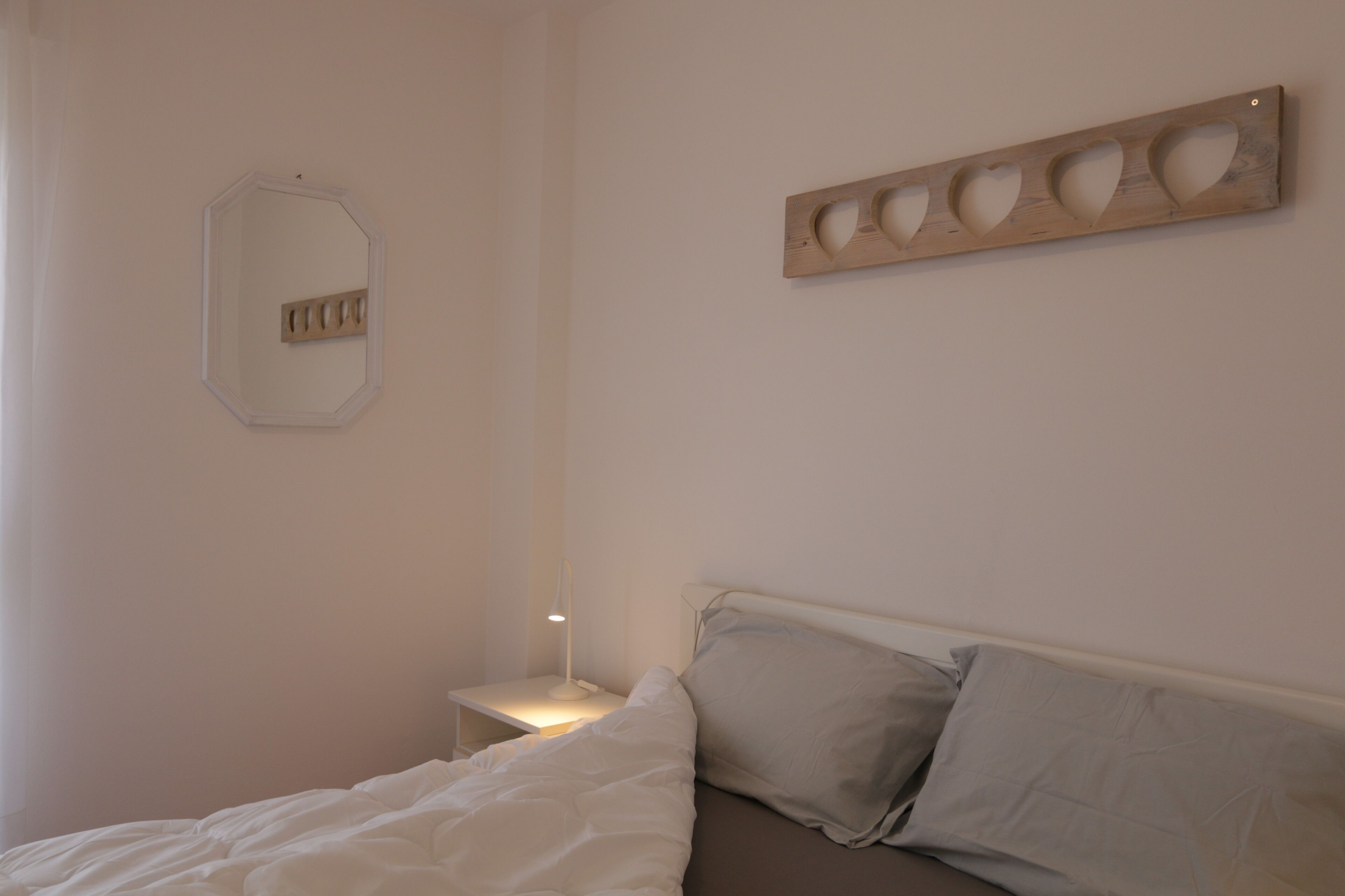 A bedroom with a bed, a bedside table with a lamp, a wall-mounted mirror, and a wooden art piece.