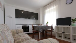 A living room with a sofa, a dining table with chairs, a kitchen with cabinets, a television on a stand, and a clock on the wall.