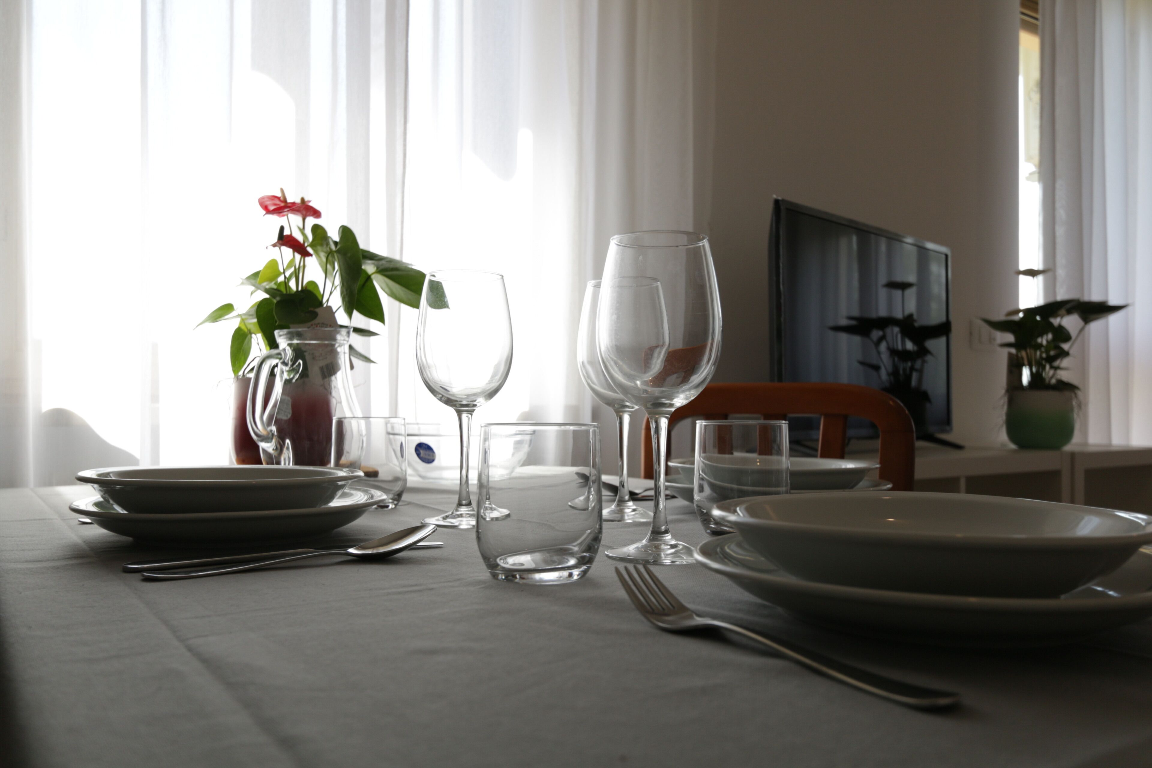 Una mesa de comedor dispuesta con platos, cubiertos y copas de vino, junto con un florero y una jarra de agua.