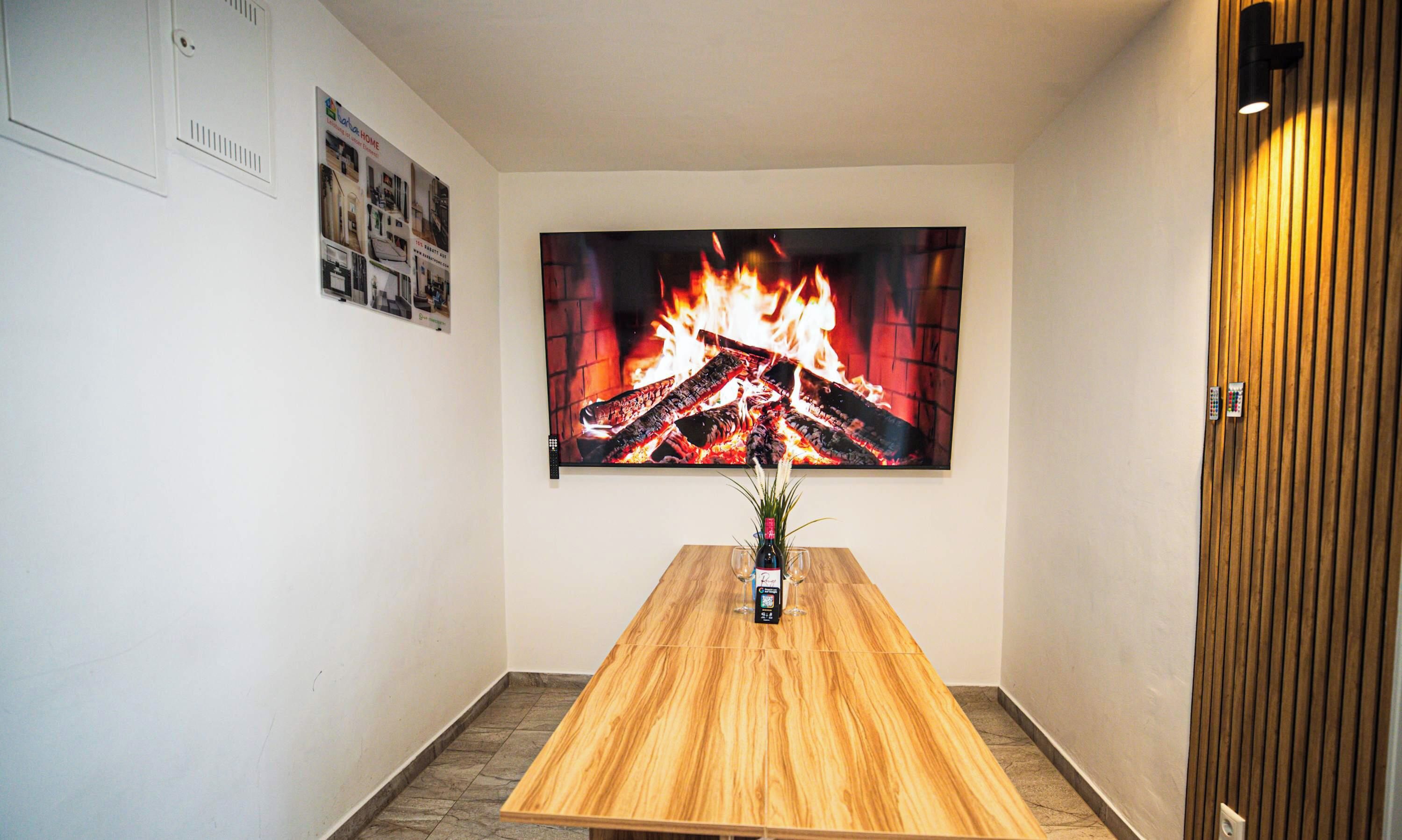 Une pièce avec une table en bois, un téléviseur fixé au mur qui diffuse une image de cheminée, et une porte ornée d’un panneau de bois vertical.