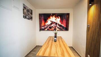 Une pièce avec une table en bois, un téléviseur fixé au mur qui diffuse une image de cheminée, et une porte ornée d’un panneau de bois vertical.