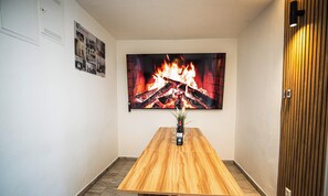 Ein Zimmer mit einem Holztisch, einem an der Wand befestigten Fernseher, der einen Kamin zeigt, und einer Tür mit einer vertikalen Holztäfelung.