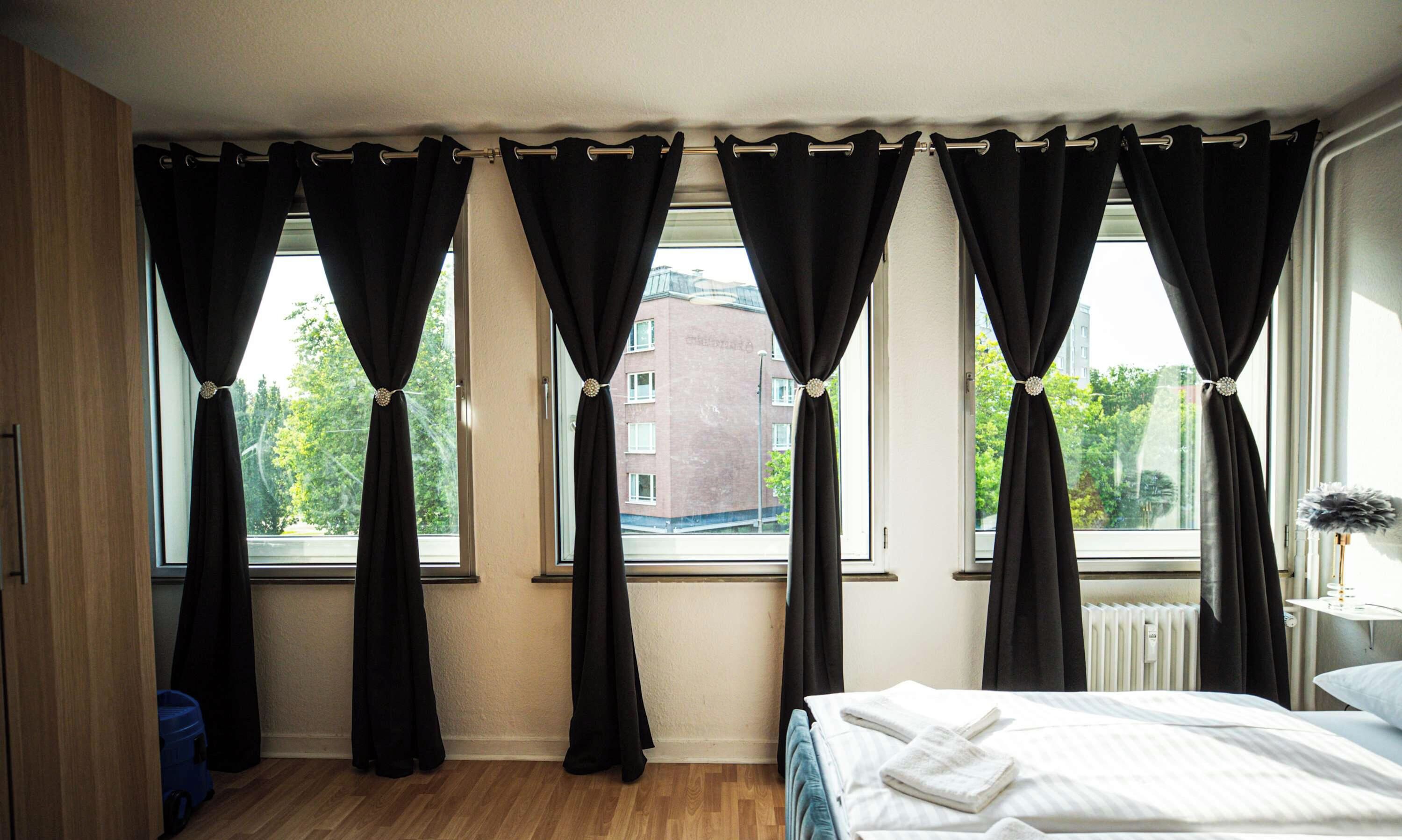 A hotel room with a bed, a window with curtains, a radiator, and a view of buildings outside.
