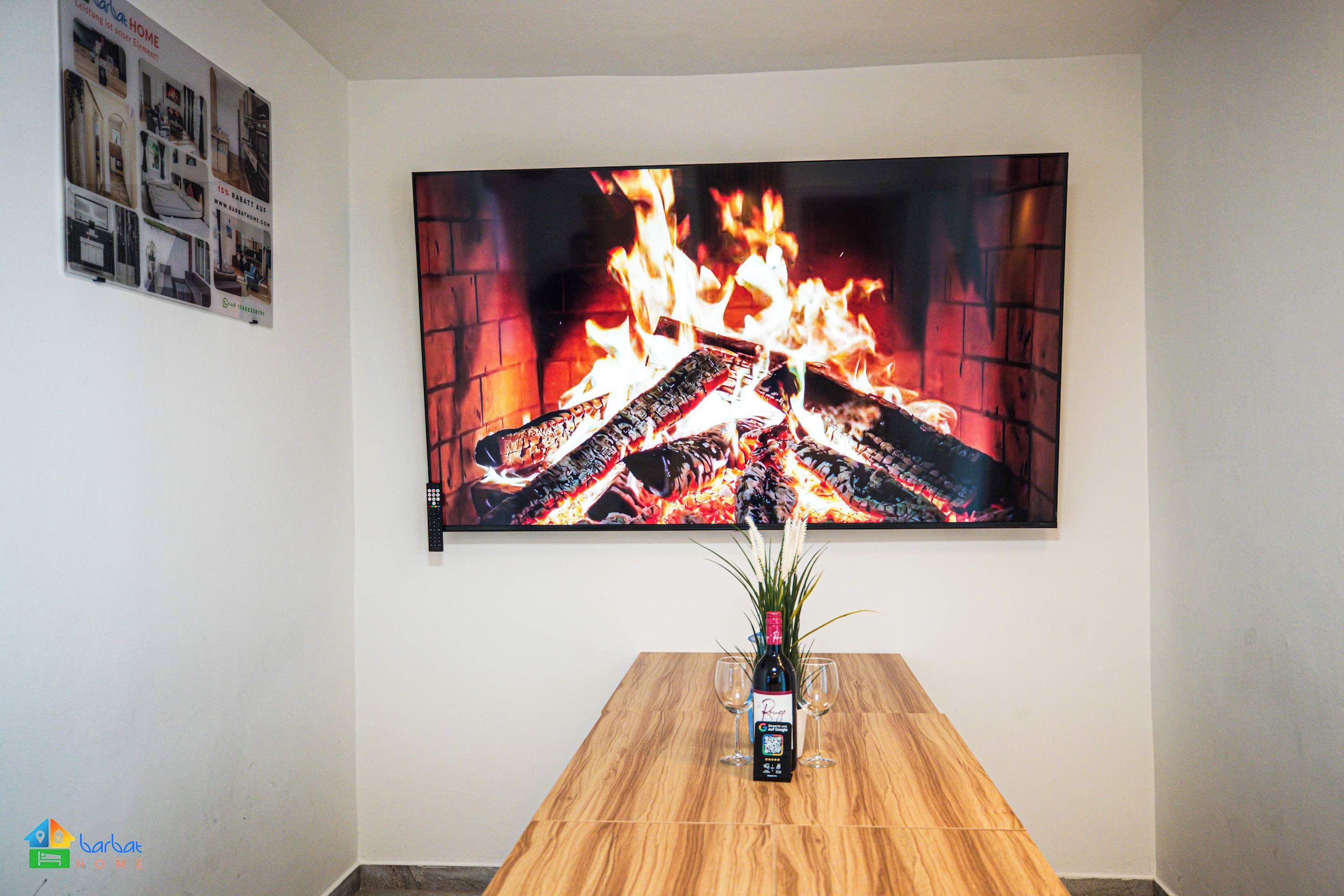 Une pièce avec un grand téléviseur à écran plat diffusant une image de feu, une table à manger en bois avec une bouteille et des verres, et une petite plante en pot.