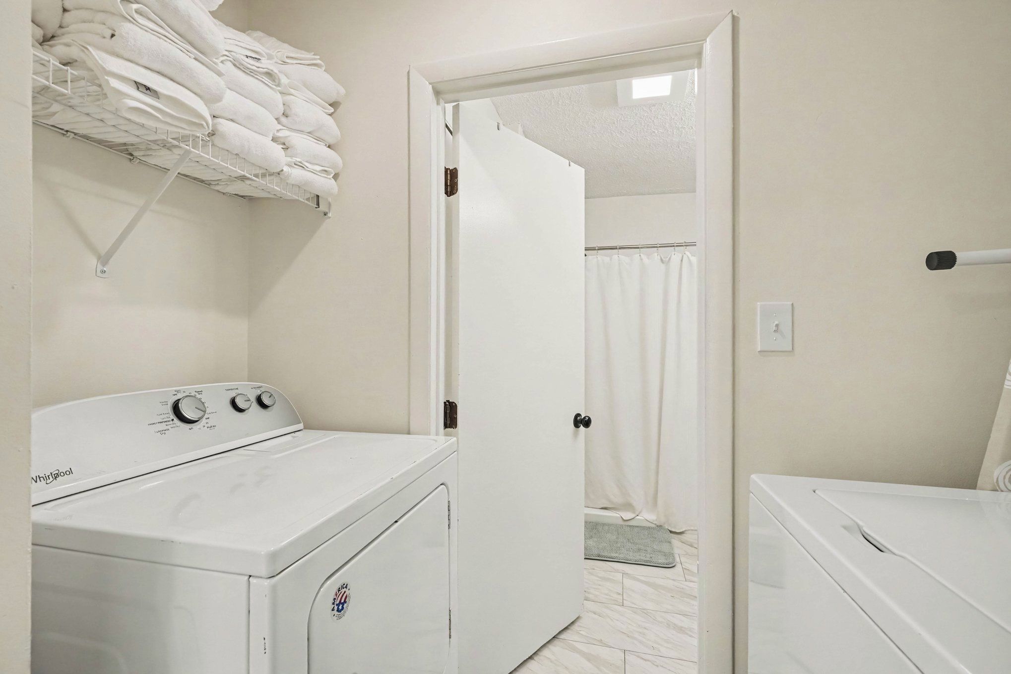 A laundry room with a front-loading washer and dryer, a towel rack above the washer, and a door leading to a bathroom with a shower curtain.