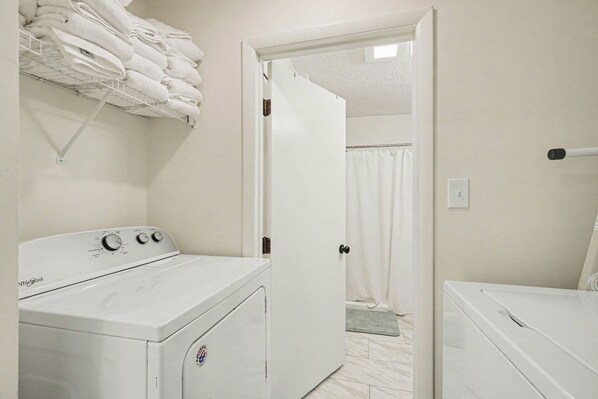 A laundry room with a front-loading washer and dryer, a towel rack above the washer, and a door leading to a bathroom with a shower curtain.