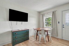 A room with a flat-screen TV mounted on the wall, a wooden dining table with white chairs, a teal dresser with drawers, and a door with a window view.