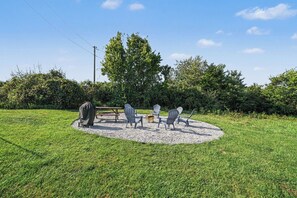 An outdoor seating area with a picnic table and chairs, surrounded by a gravel circle, with a grassy field and trees in the background.