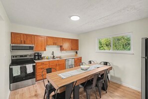 A kitchen with wooden cabinets, a stainless steel oven, a dining table with chairs, and a window with a view of trees.