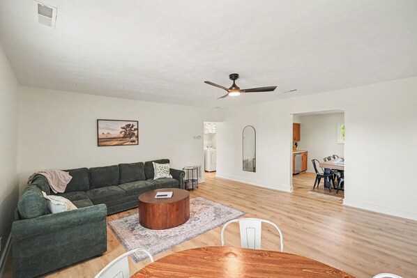 A modern living room with a green sofa, a round coffee table, and a dining area with a wooden table and chairs.