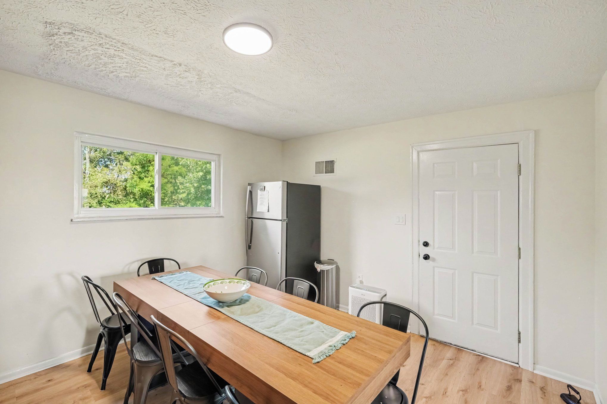 A compact kitchen with a dining table, chairs, a refrigerator, and a window with a view of greenery.