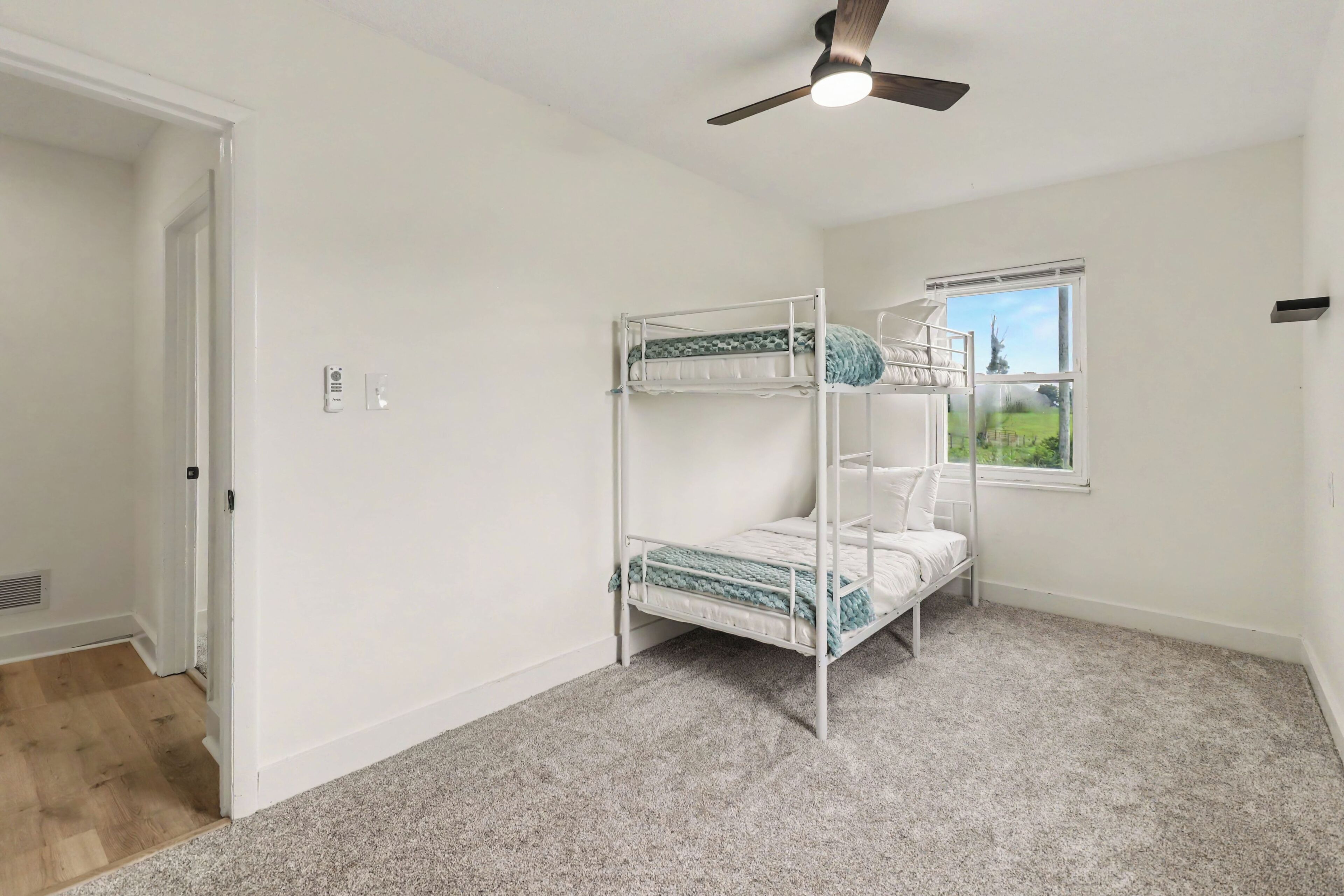 A room with a bunk bed, a ceiling fan, a window with a view, and a door.