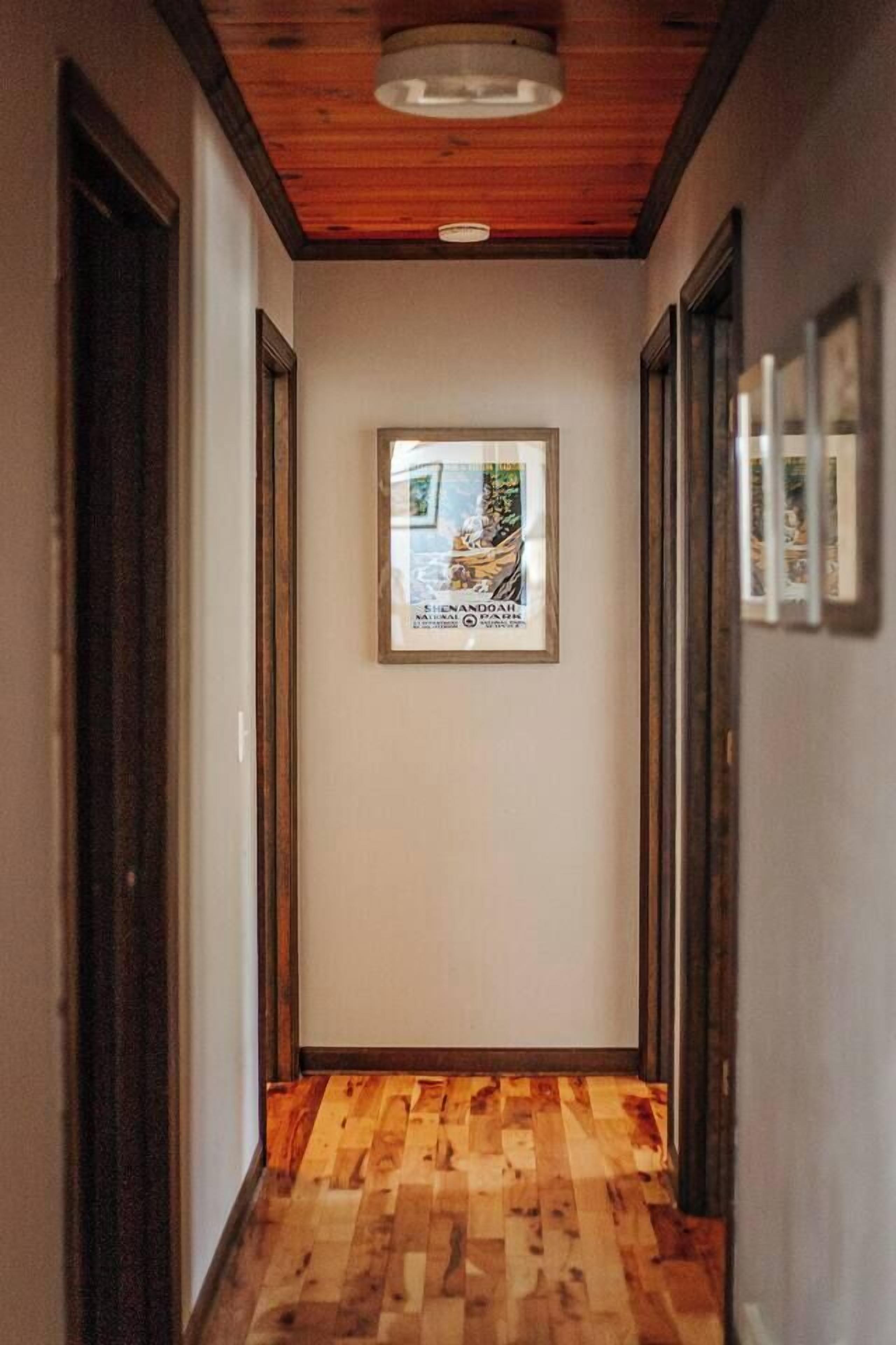 A hallway with wooden flooring, framed artwork on the wall, and wooden doors.