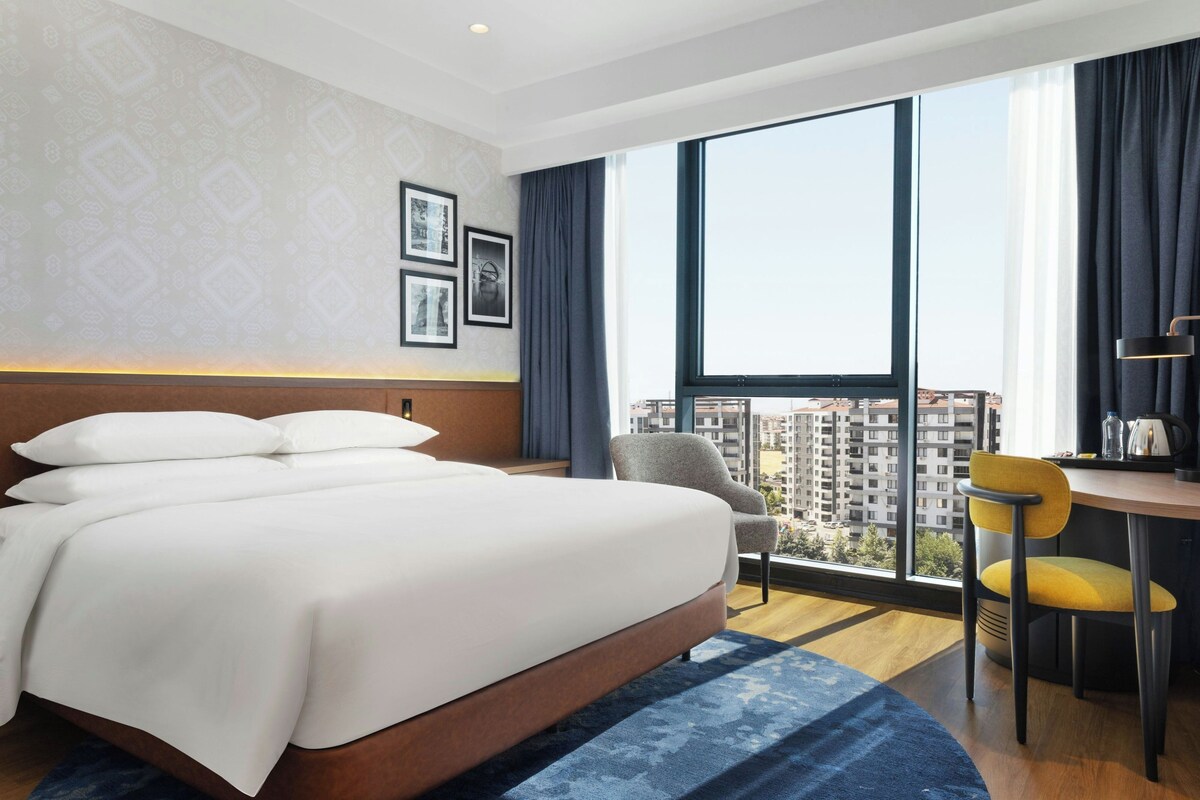 Habitación de hotel con una cama grande, un escritorio con silla, vista a edificios por la ventana y un papel tapiz estampado.