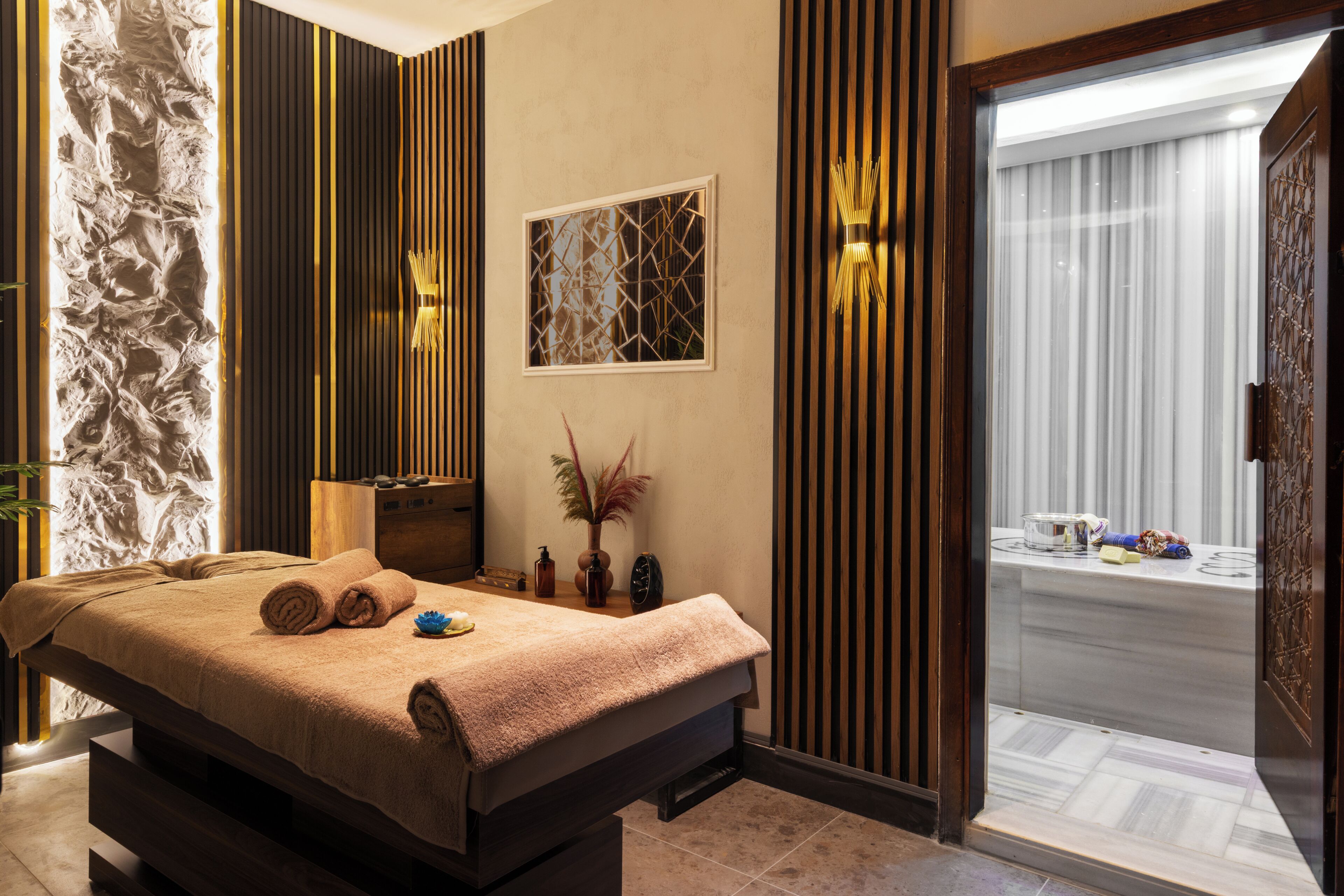 A spa room with a massage table, a stone wall, wooden panels, a window with a view, and a door leading to a bathroom.